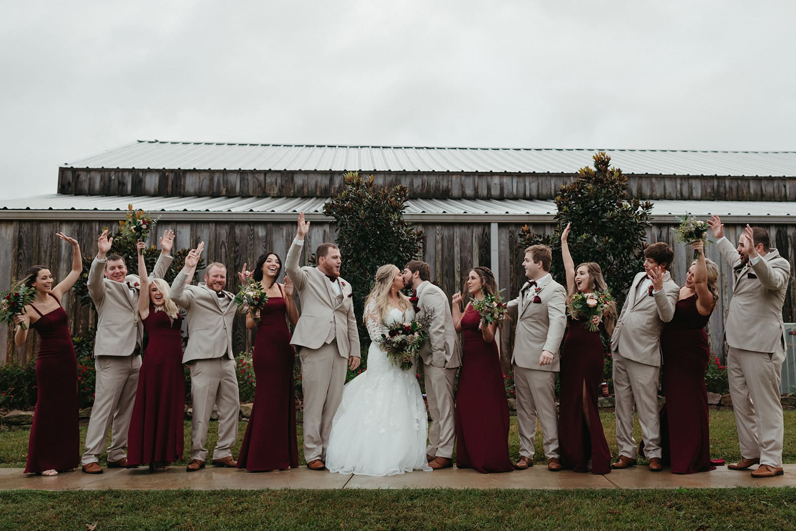 A wedding party stands in front of a rustic wooden building, with the bride and groom kissing in the center. Bridesmaids in burgundy dresses and groomsmen in beige suits raise their arms and smile joyfully.