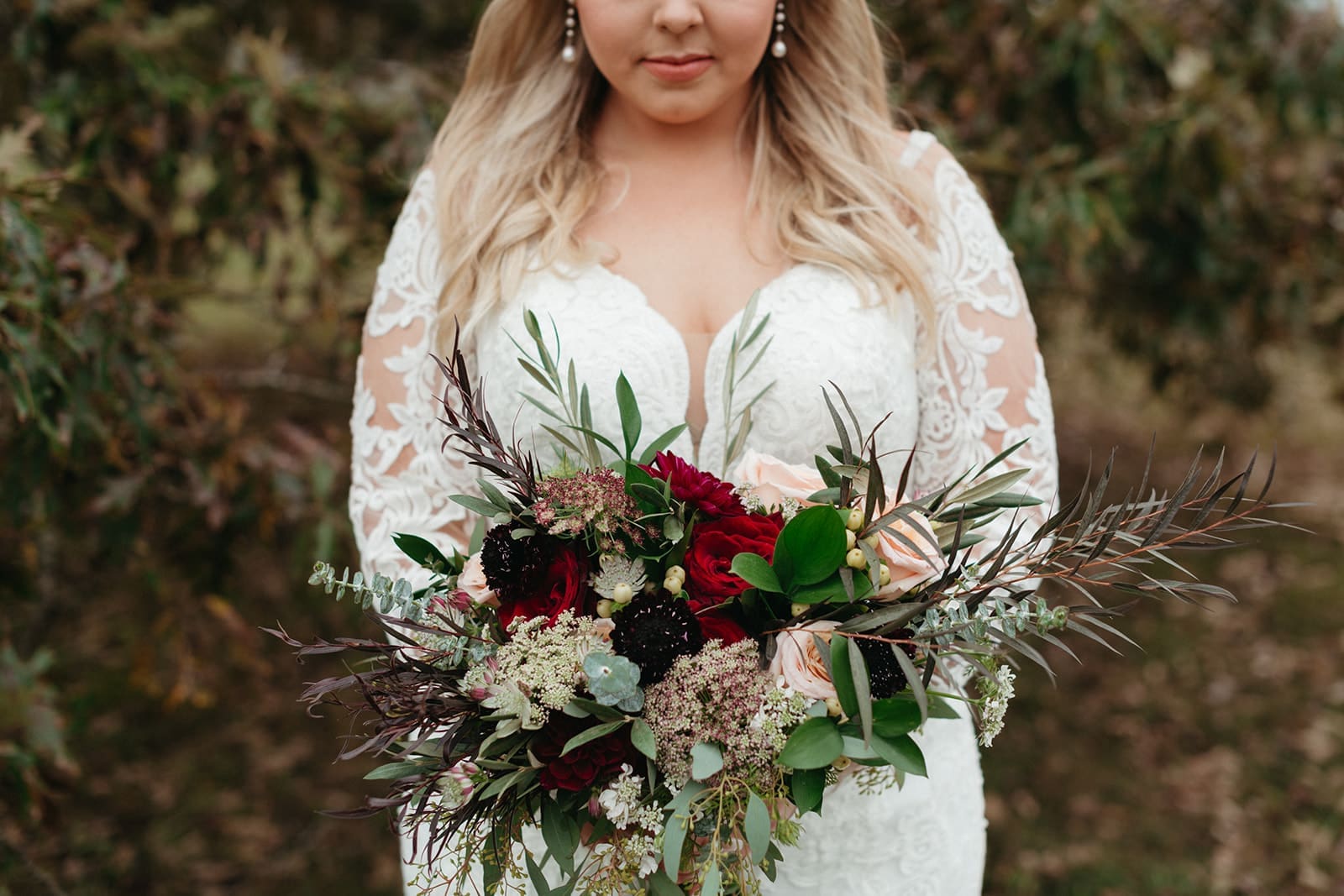 A bride in a lace wedding dress holds a vibrant bouquet with red, pink, and dark flowers, and green foliage, outdoors with greenery in the background. Her face is partially visible.