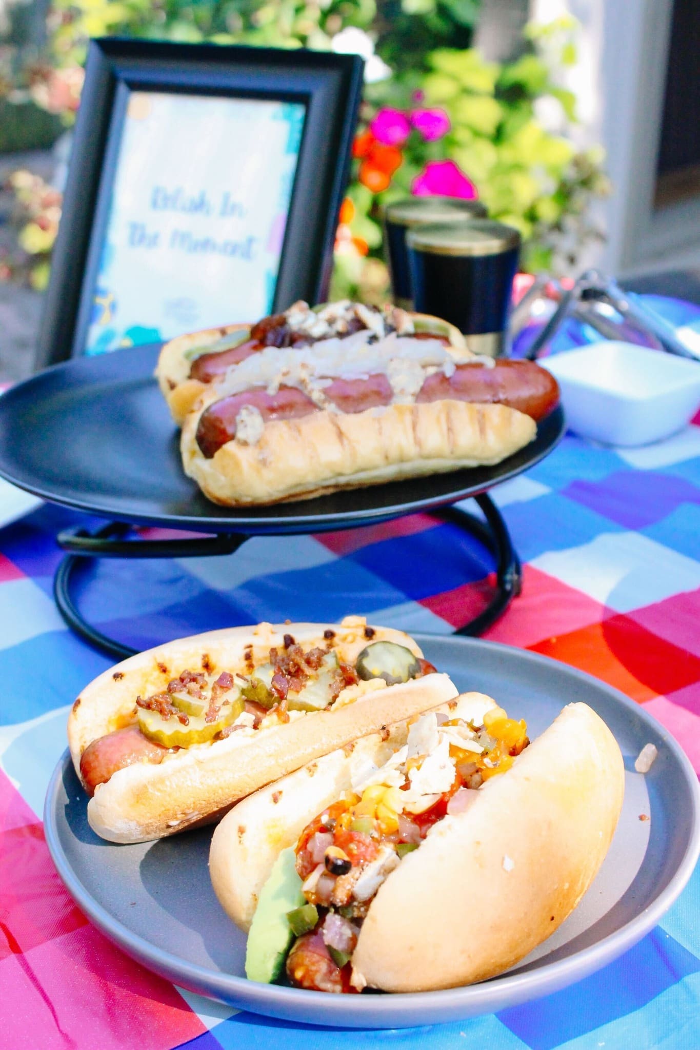 Three gourmet hot dogs with assorted toppings are displayed on plates atop a colorful checkered tablecloth at an outdoor gathering. A framed sign and flowers are blurred in the background.