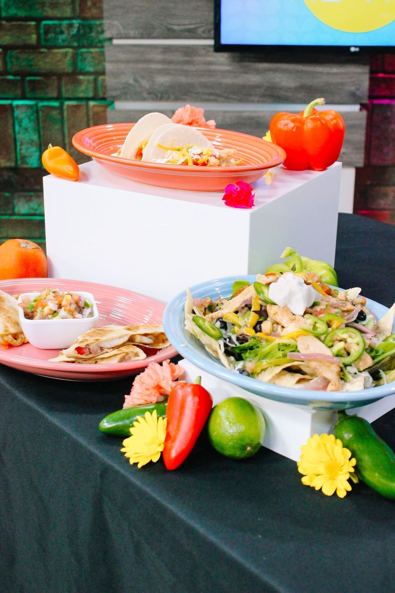 Colorful plates of Mexican food, including nachos with sour cream, tacos, salsa, and assorted bell peppers, limes, and flowers arranged on a black table with a white display stand.
