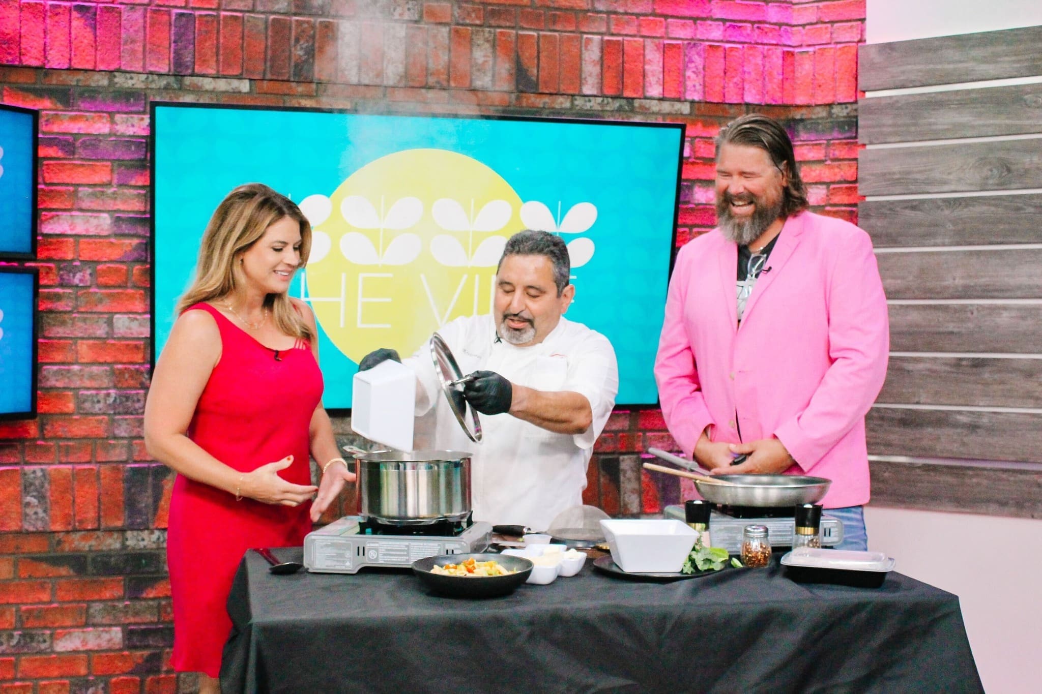 Three people stand around a cooking station on a TV set. A chef stirs a steaming pot, while a woman in a red dress and a man in a pink blazer smile and watch. Cooking utensils and food are on the table.