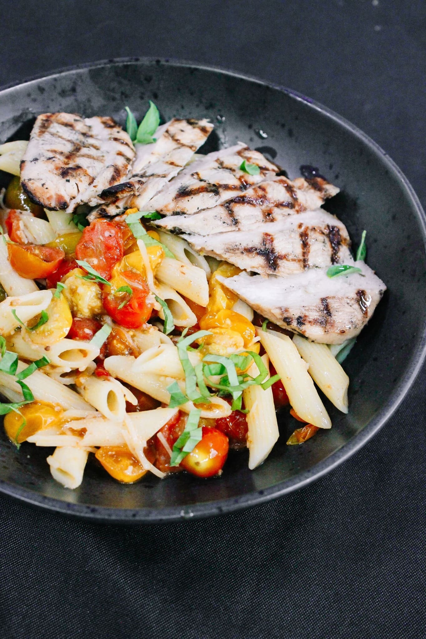 A black bowl filled with penne pasta, cherry tomatoes, and grilled chicken slices, garnished with fresh herbs, on a dark background.