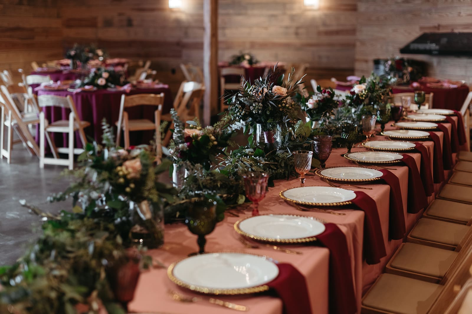 A long table set for a formal event with white plates, gold-rimmed chargers, pink and gold tablecloths, and floral centerpieces. Wooden chairs and softly lit wooden walls create a warm, rustic atmosphere.