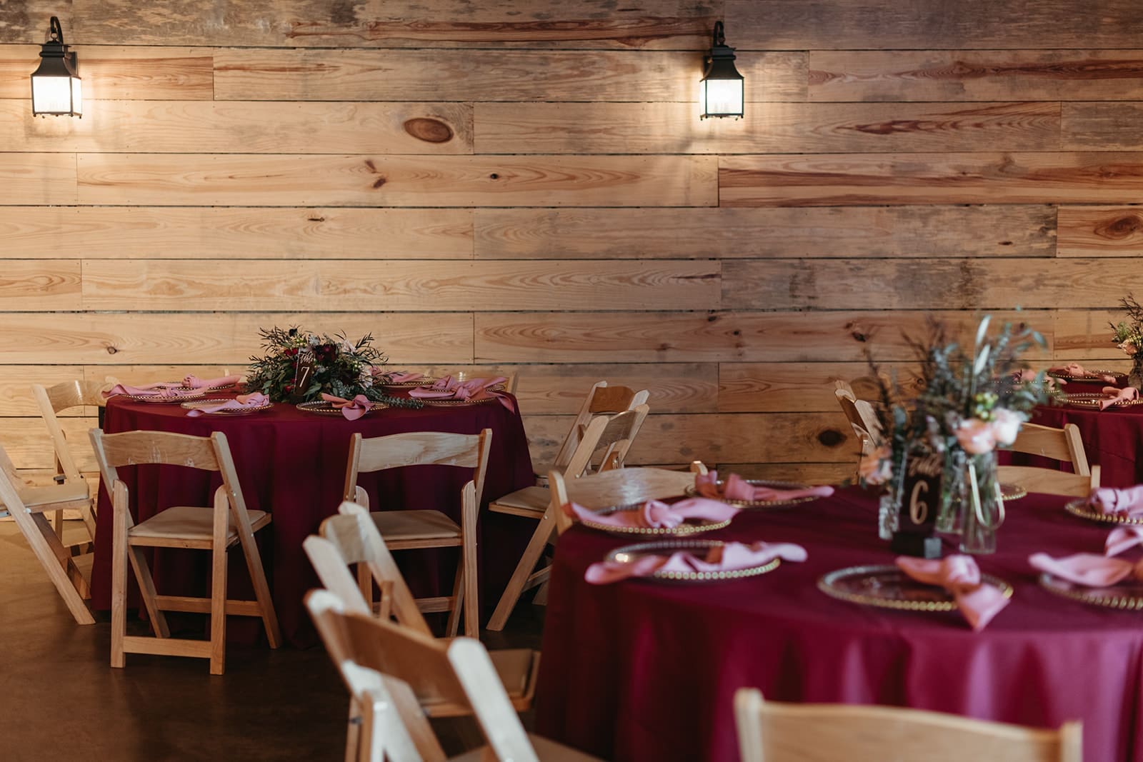 Round tables covered with deep red tablecloths are set for an event, with wooden chairs, pink napkins, and floral centerpieces. The room features wooden plank walls and soft wall-mounted lighting.