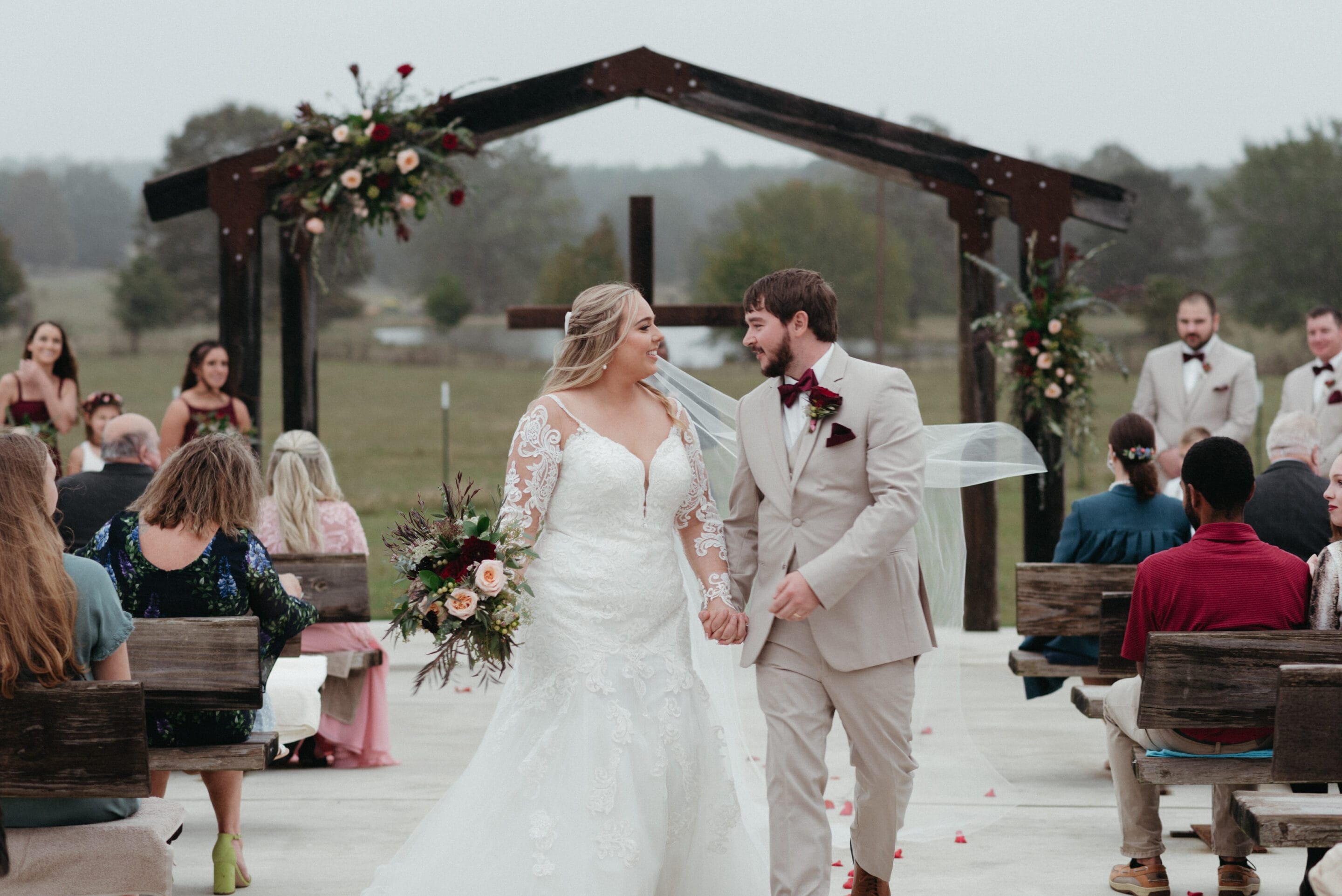 A bride and groom walk hand-in-hand down the aisle outdoors, smiling at each other. The bride wears a lace wedding dress and holds a bouquet. Guests sit on benches, and a decorated wooden arch stands behind them.