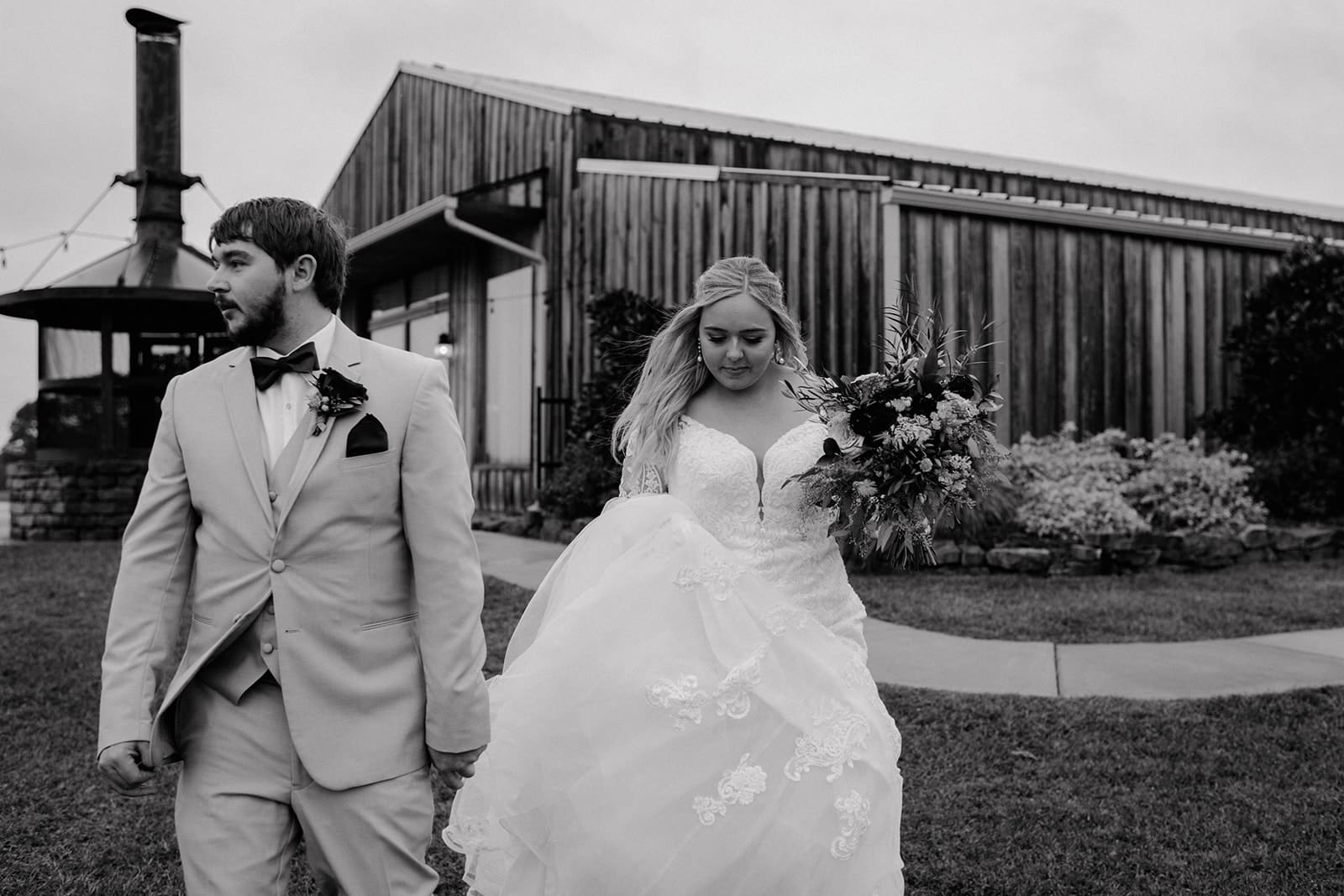 A bride and groom walk together outside a rustic wooden building. The groom wears a light suit with a bow tie, and the bride holds her floral bouquet and dress, both looking thoughtful. The scene is in black and white.