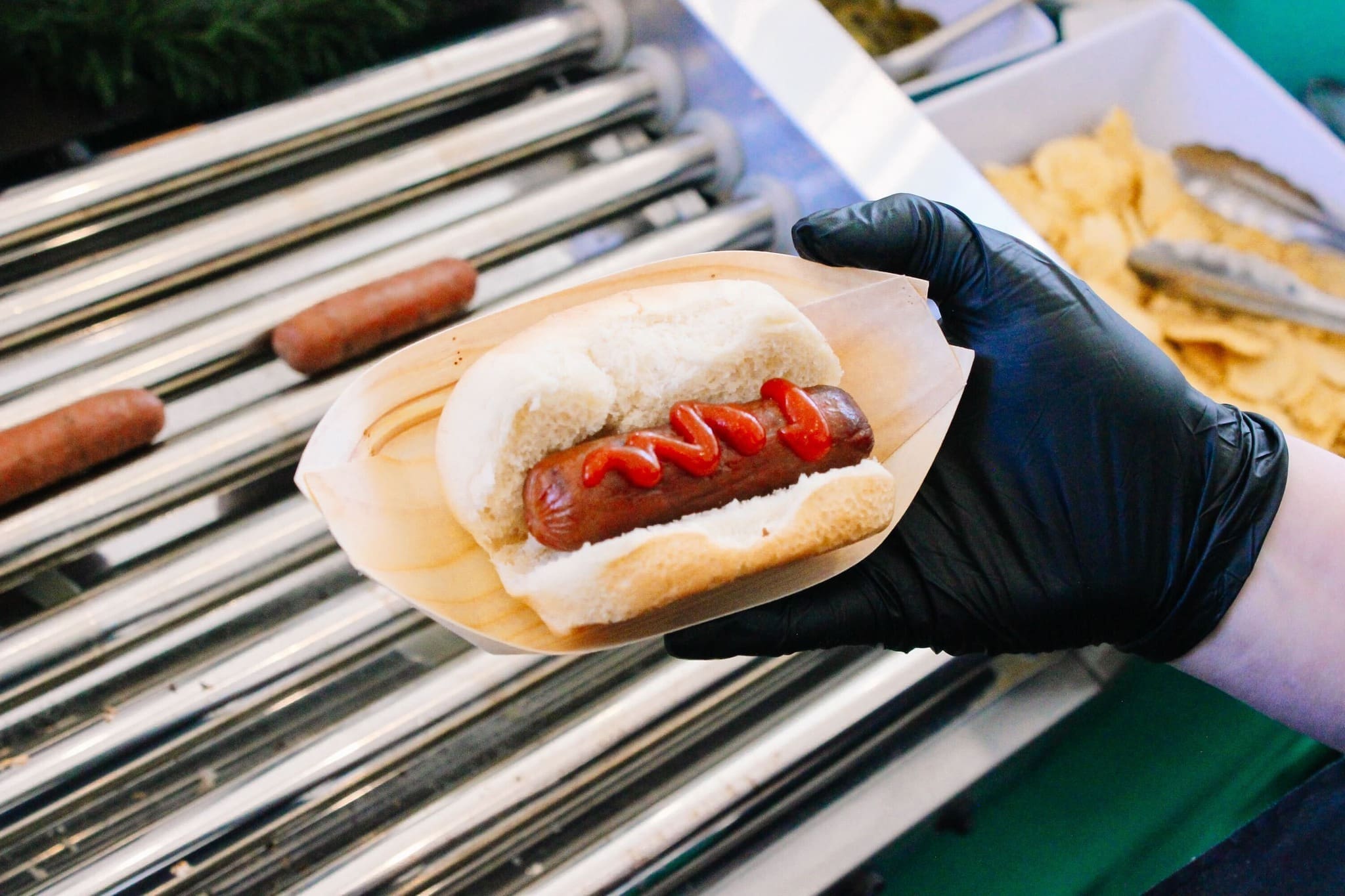 A person wearing a black glove holds a hot dog with ketchup in a bun. Hot dogs are grilling in the background, and there is a tray of chips and tongs nearby.