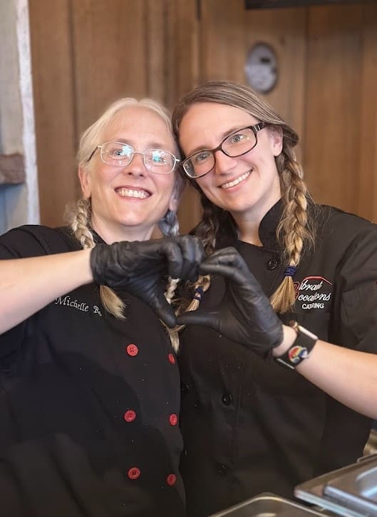 Two women with braided hair and glasses, wearing black chef jackets, smile and make a heart shape with their gloved hands together in a kitchen setting.