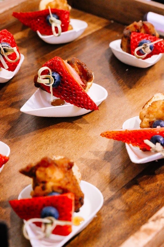 Small appetizer bites served on white dishes, each topped with a slice of strawberry, a blueberry, and skewered with a toothpick, arranged on a wooden surface.