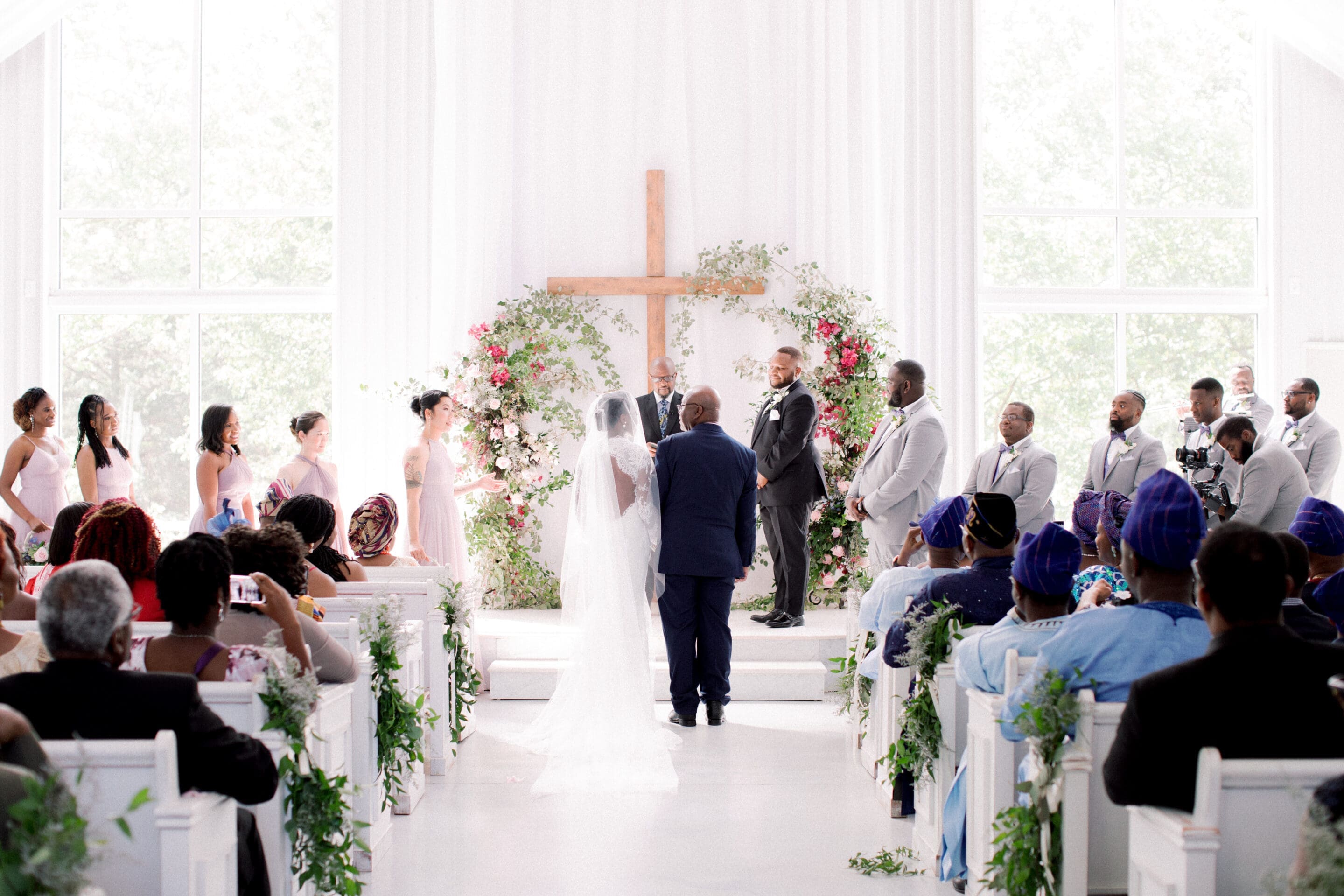 A bride and groom stand at the altar in a bright chapel with a large wooden cross and floral arch, surrounded by bridesmaids, groomsmen, and seated guests, as the bride’s escort gives her away.