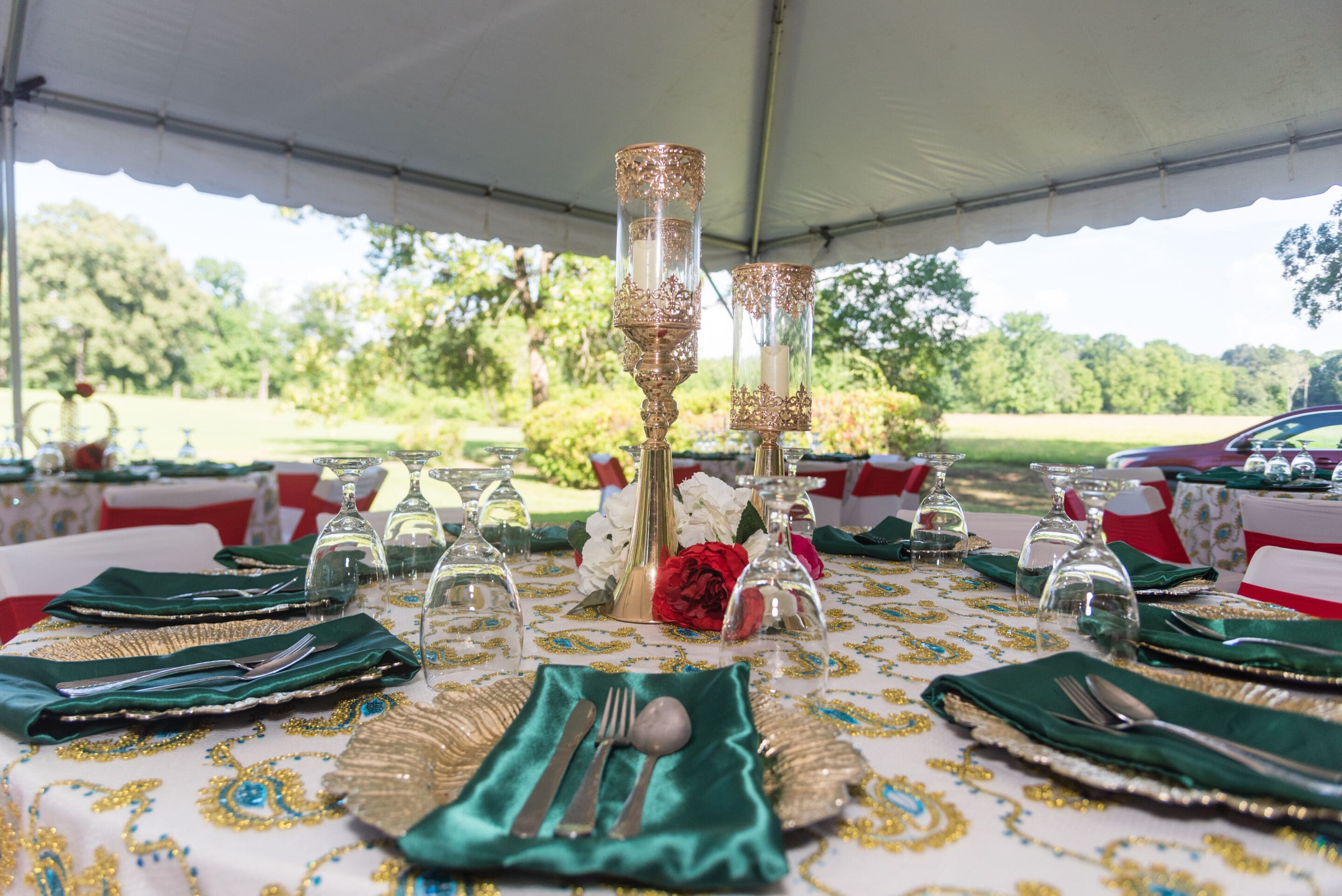A decorated outdoor table set for an event with green napkins, gold plates, glassware, and gold candle holders under a white tent, surrounded by greenery.