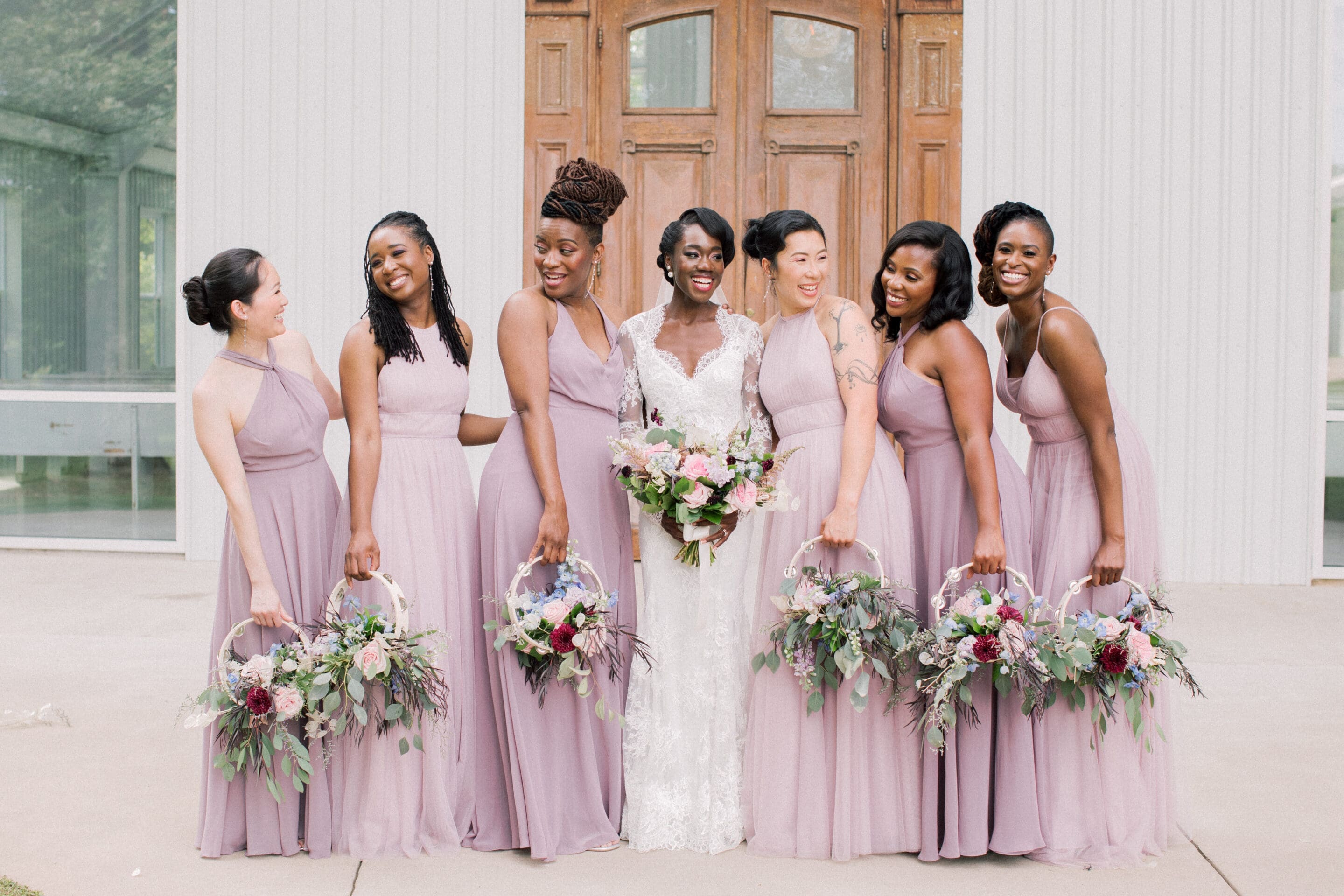 A bride in a white lace gown stands outside with six bridesmaids in matching light purple dresses, each holding a floral hoop bouquet. They are smiling and posing in front of wooden doors.