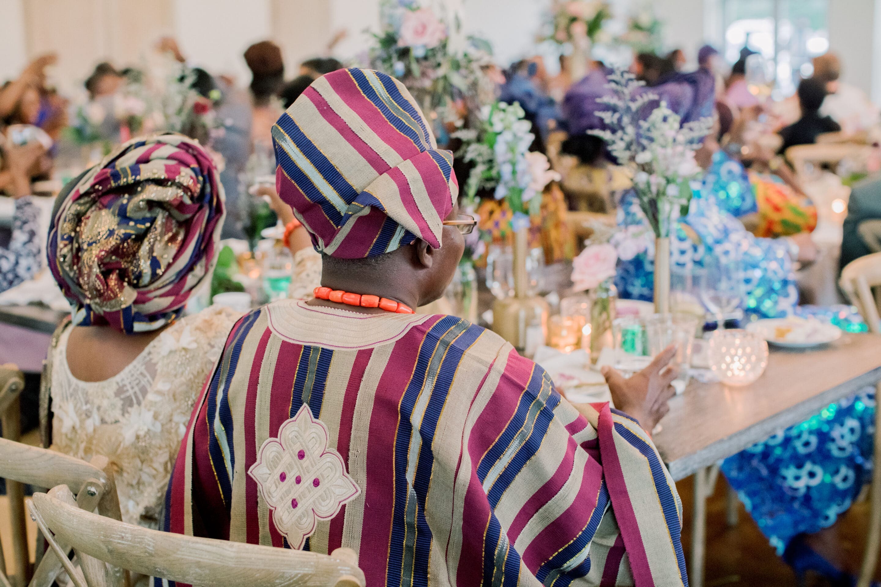 A person wearing a colorful striped traditional outfit and headwrap sits at a decorated table during a festive gathering; guests in vibrant attire and floral arrangements fill the background.