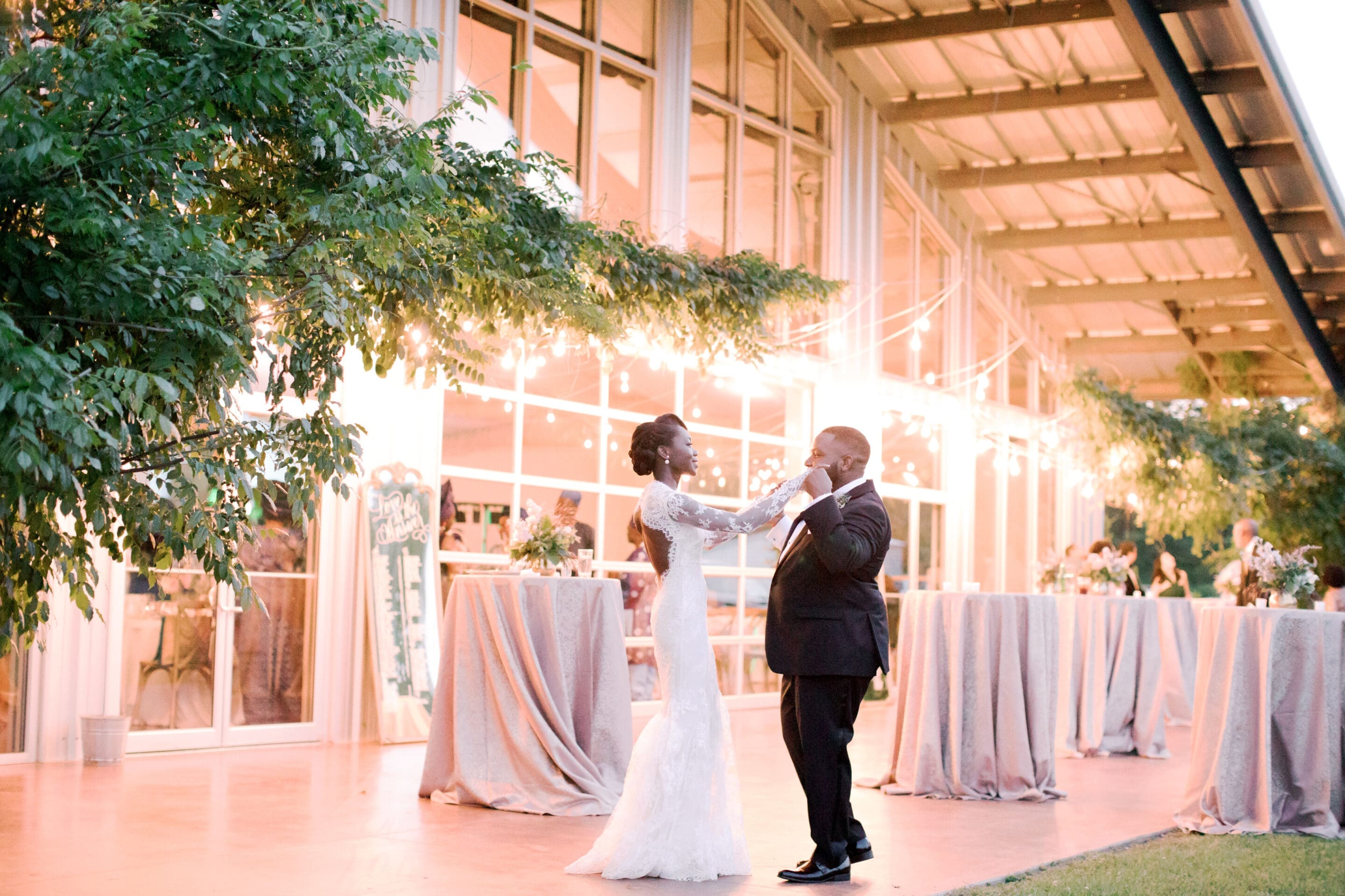 A bride and groom share a joyful first dance outside a modern venue, surrounded by greenery and softly glowing string lights. Tables covered in light cloths are set up in the background.