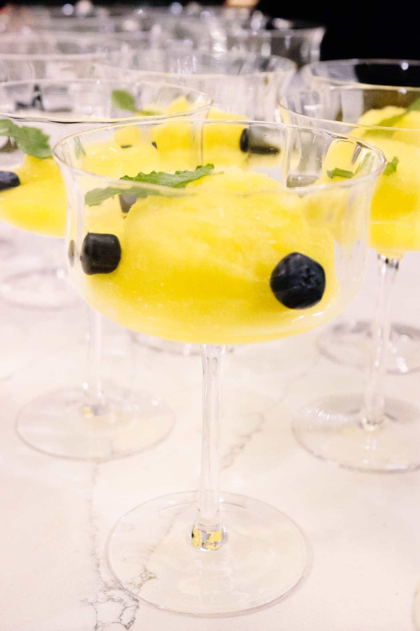 Several cocktail glasses filled with bright yellow sorbet, garnished with blueberries and a mint leaf, are arranged on a white marble surface.